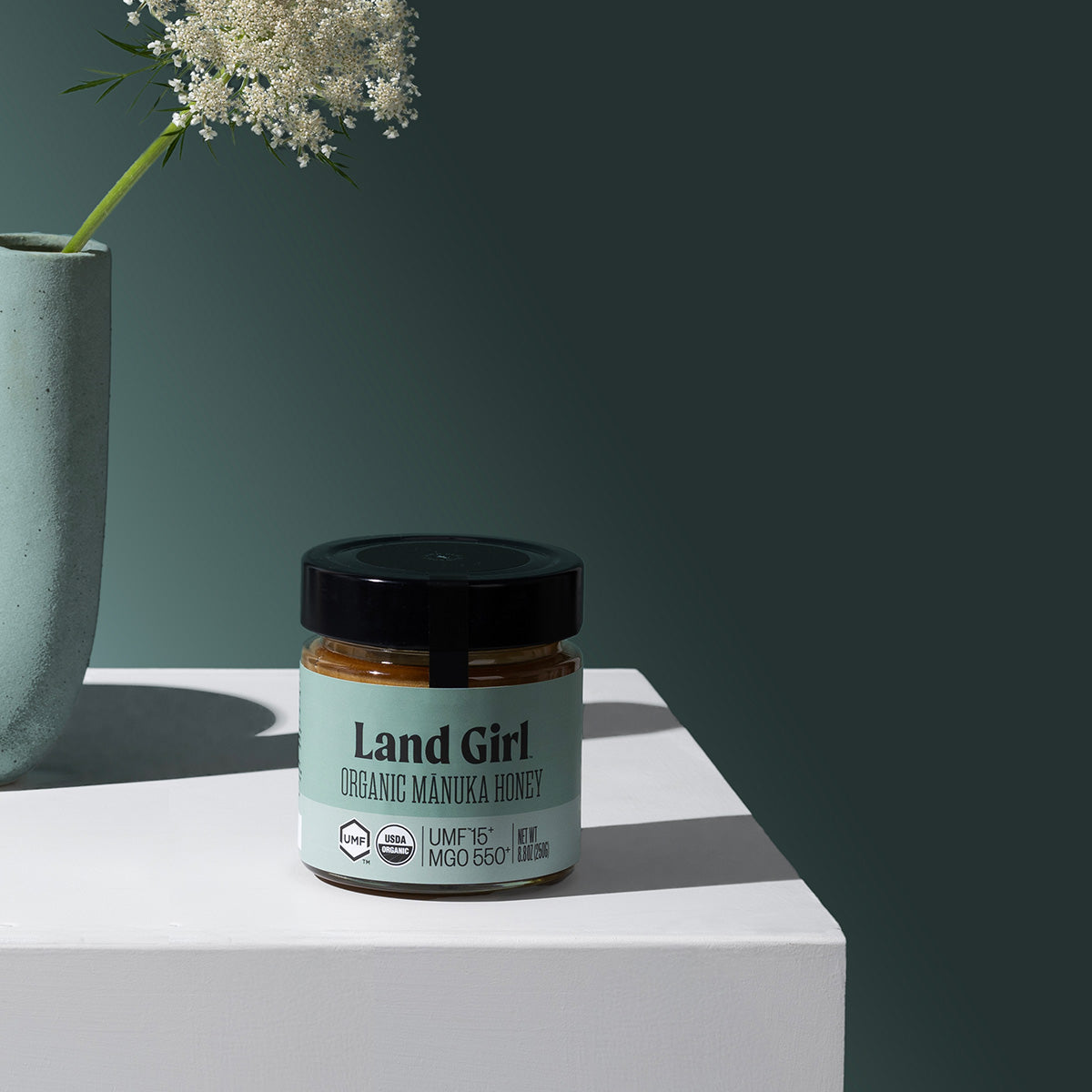 A jar of Land Girl Organic Manuka Honey sits on a white pedestal against a dark teal background, with a light blue vase of white flowers in the background.