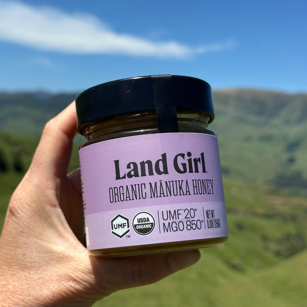 A hand holds a jar of Land Girl Organic Manuka Honey against a blurred mountain landscape background under a blue sky. 