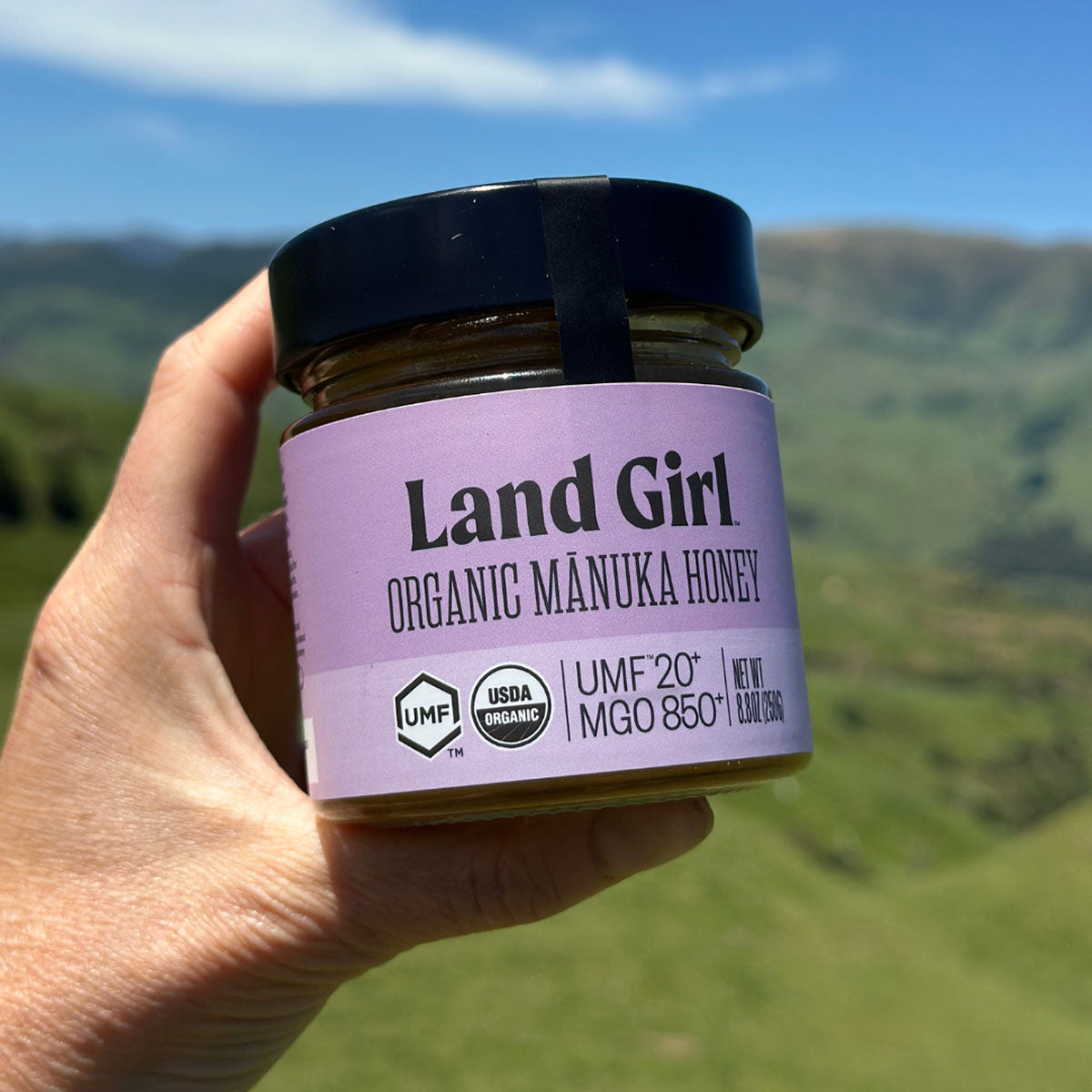 A jar of Land Girl Organic Mânuka Honey UMF20+ against a blurred green mountain landscape background. 
