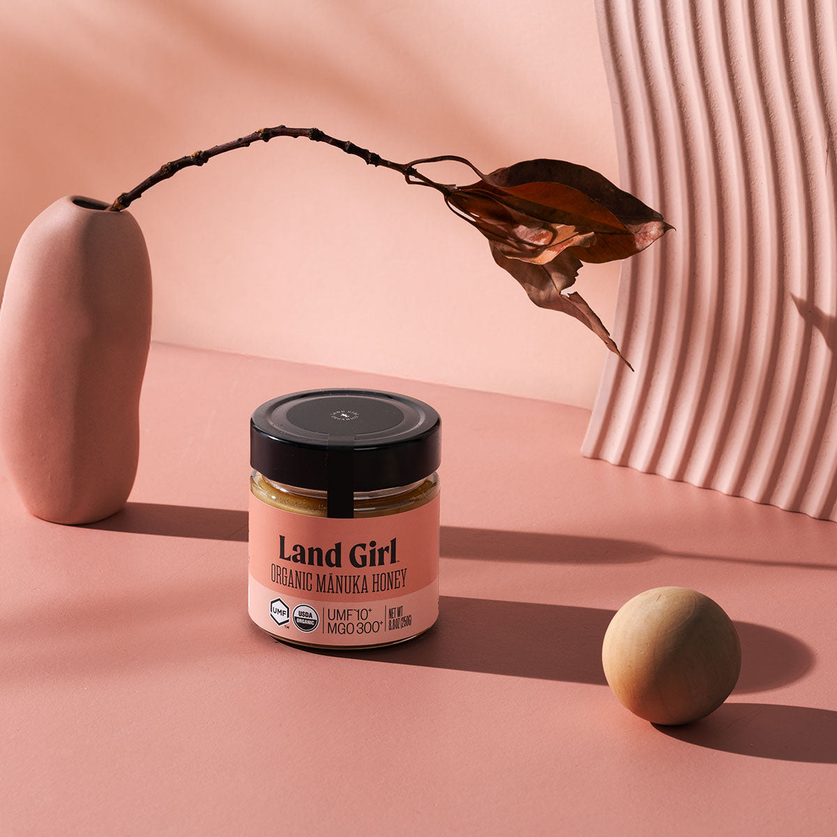 A jar of UMF10+ Land Girl Organic Manuka Honey sits on a pink surface with a pink vase holding a dried leaf.