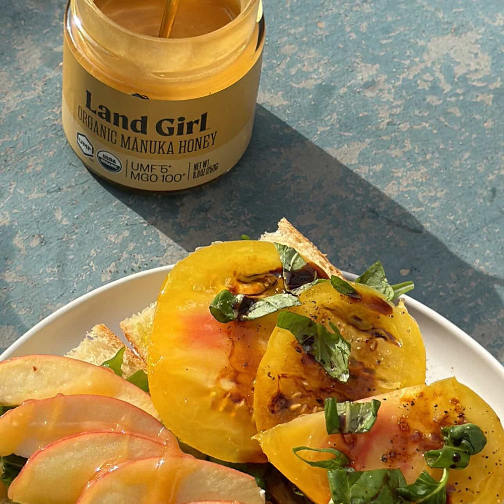 A jar of Land Girl Organic Mānuka Honey next to a plate of honey-sweetened fruit slices.