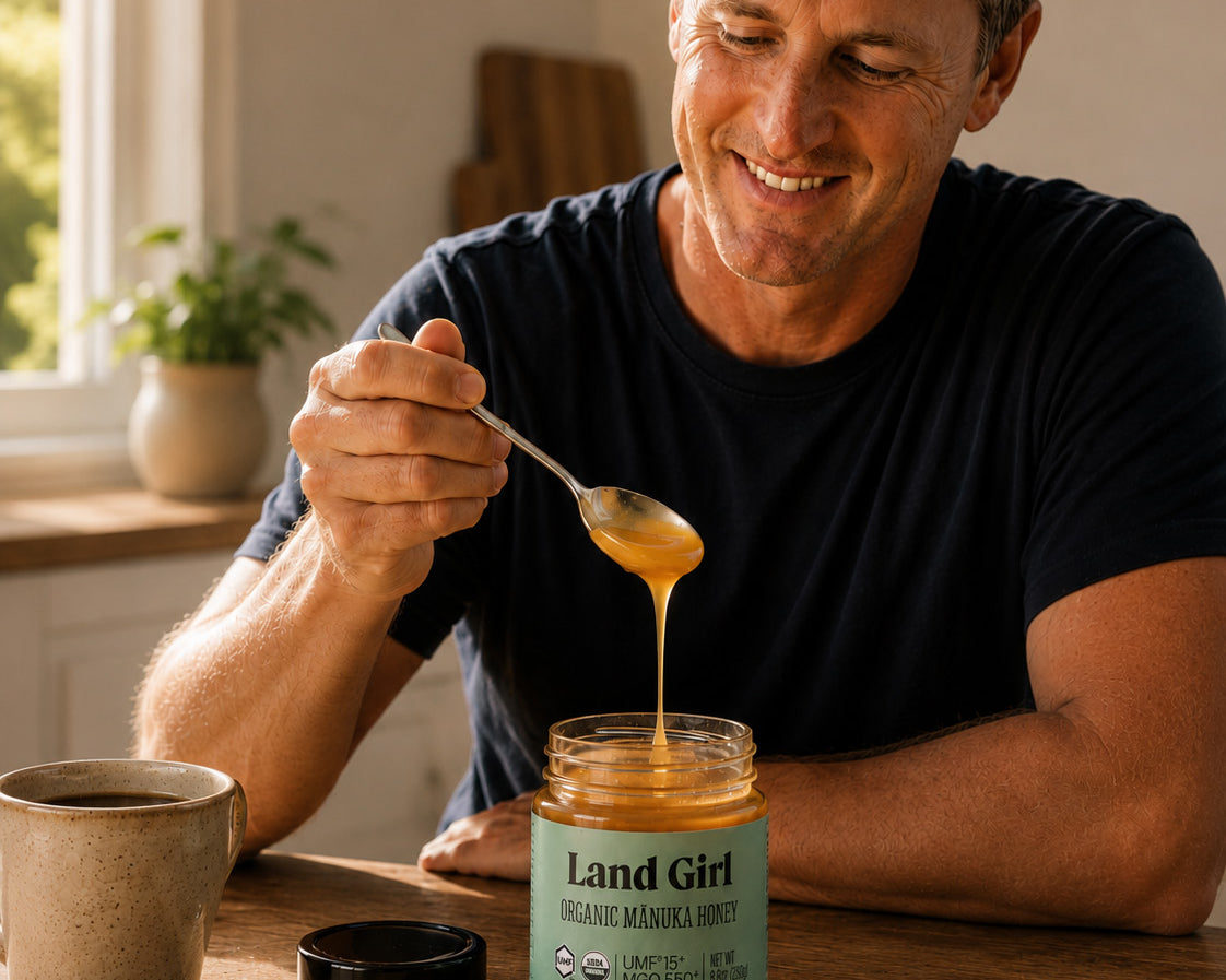 Man pouring honey from a jar labeled 'Land Girl' into a spoon, with a jar of honey and a cup on a wooden table.