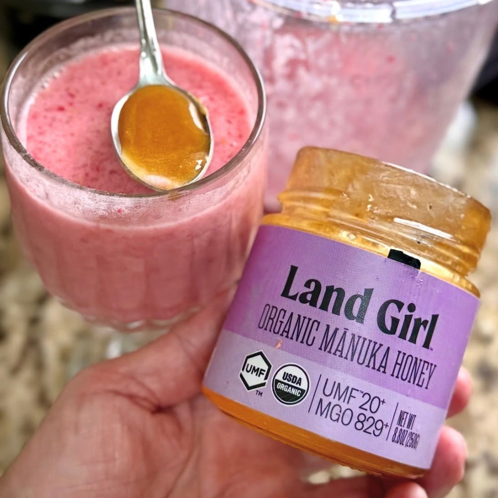 Jar of Land Girl Organic Manuka Honey with a pink smoothie and honey spoon