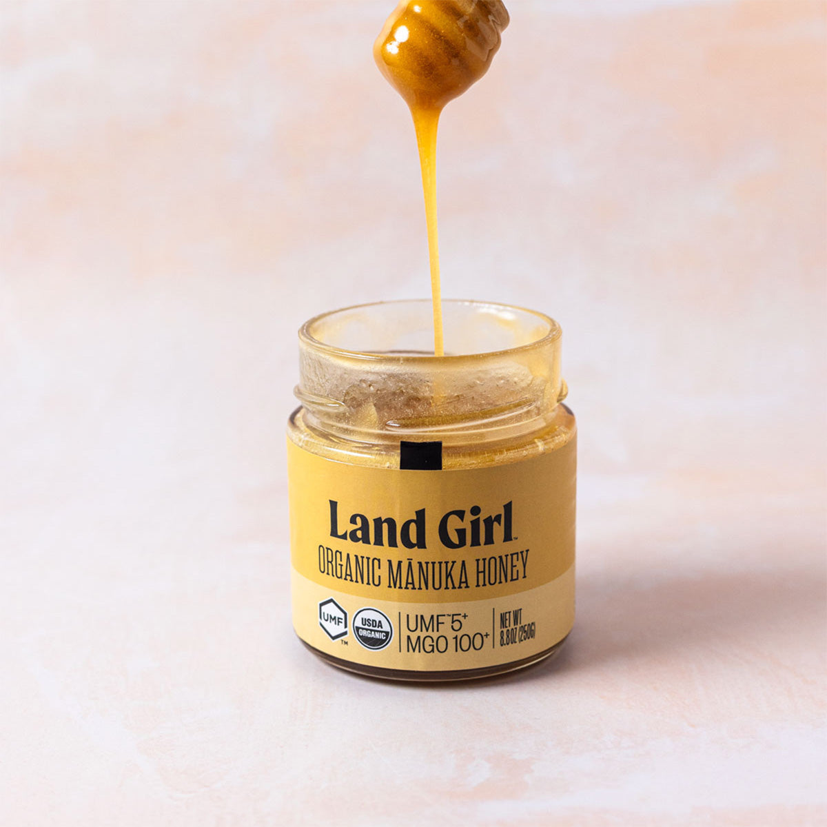 An open glass jar of Land Girl Organic Mānuka Honey with honey dripping from a wooden dipper above it, set against a soft, light beige background.