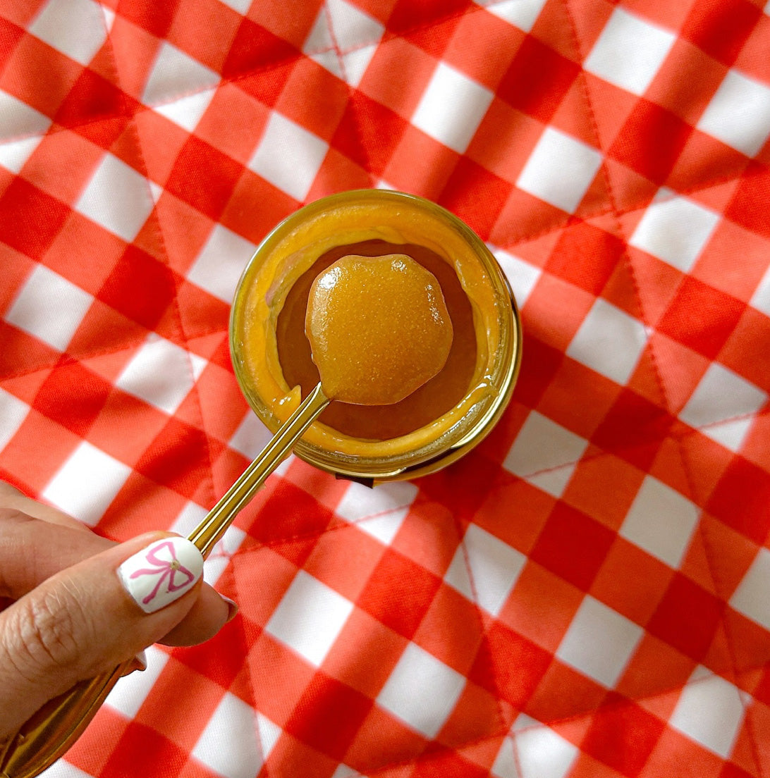 A gold spoon with a dollop of golden-organic mānuka honey over an open glass jar, set against a red and white checkered quilted blanket.